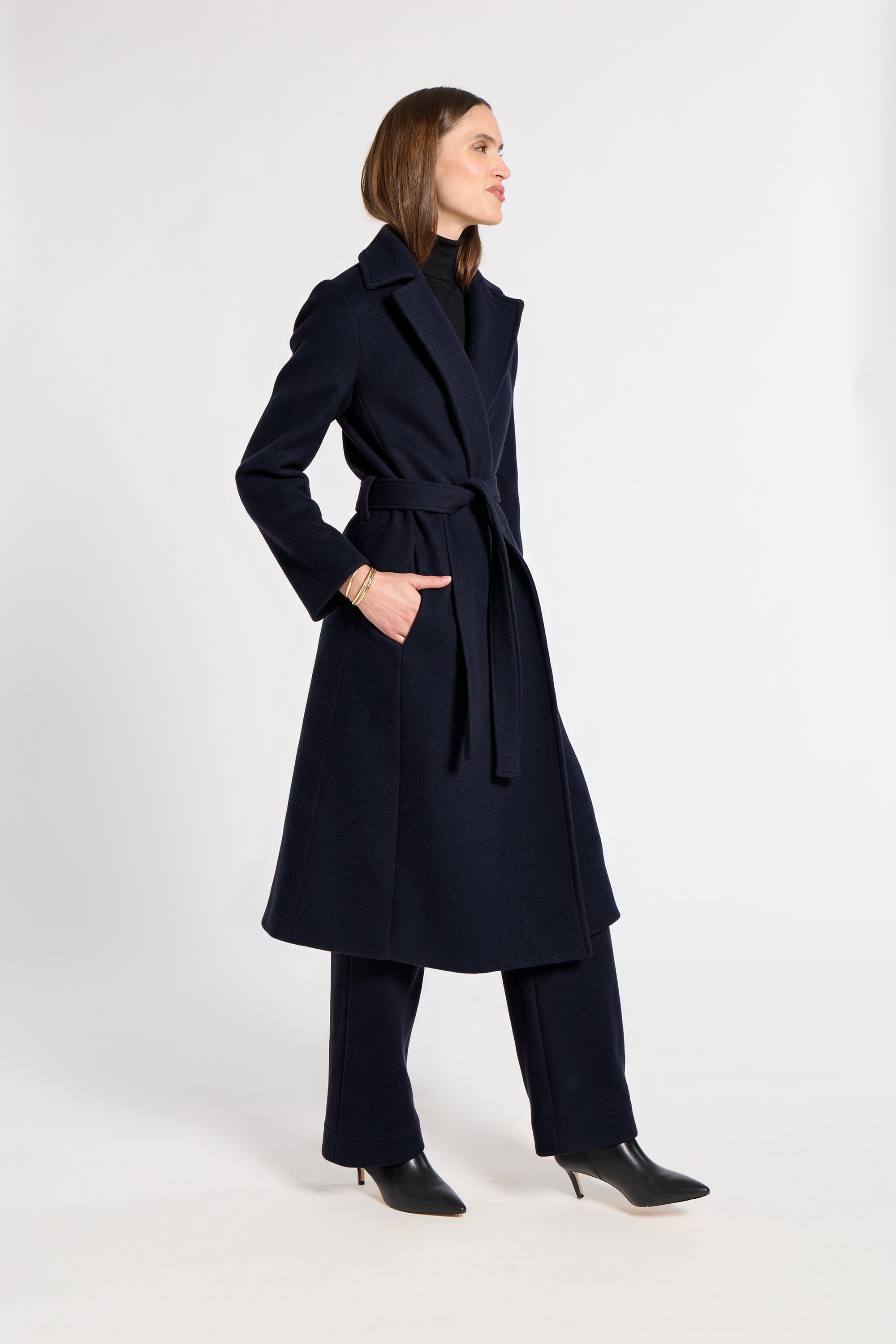 A woman modeling a long navy blue belted cashmere coat over matching navy trousers and black pointed-toe heels, standing against a plain light gray studio background.