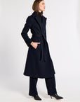 A woman modeling a long navy blue belted cashmere coat over matching navy trousers and black pointed-toe heels, standing against a plain light gray studio background.