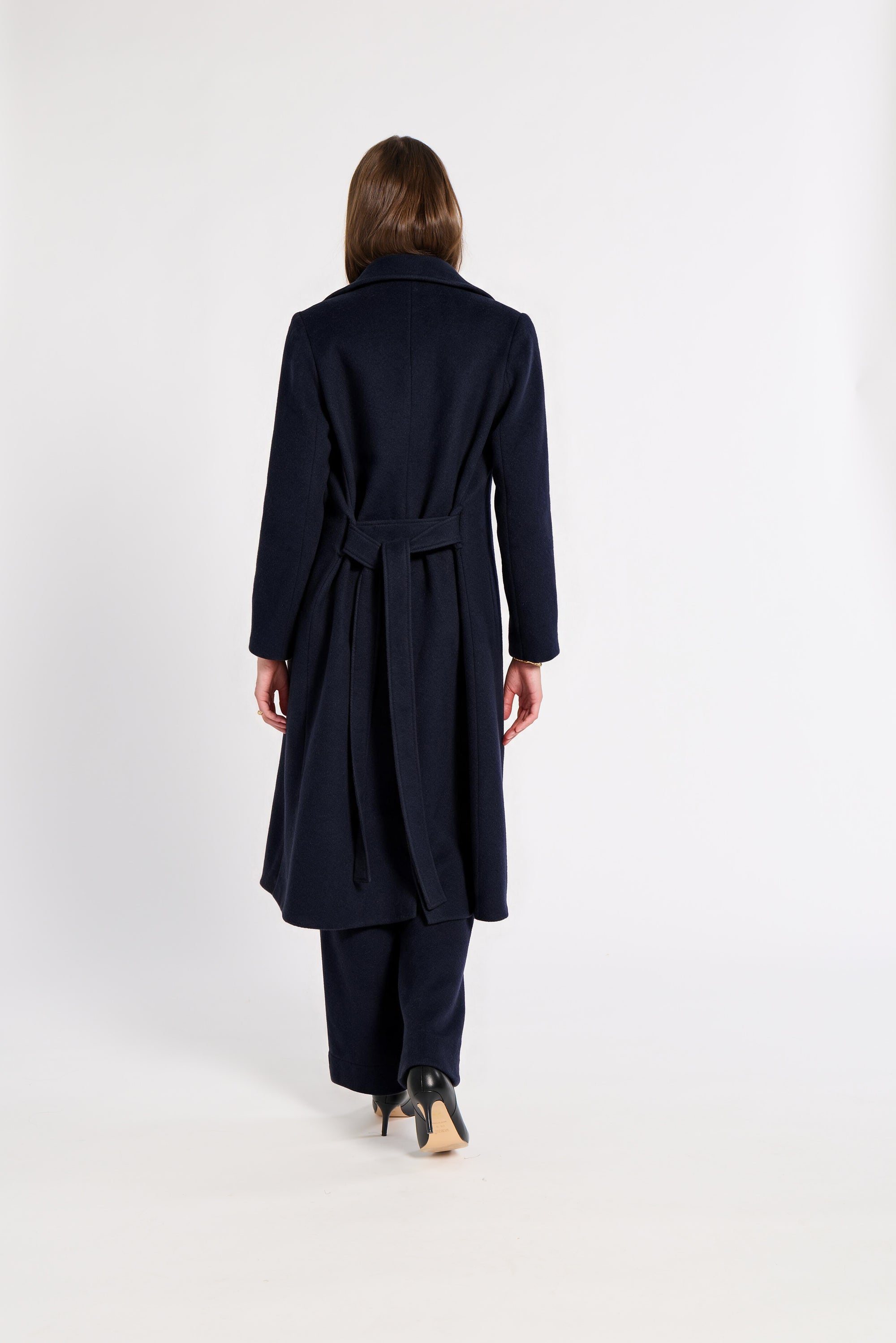 A woman viewed from behind wearing a long navy belted coat, black pants, and black high-heeled shoes against a plain white studio background.