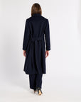 A woman viewed from behind wearing a long navy belted coat, black pants, and black high-heeled shoes against a plain white studio background.