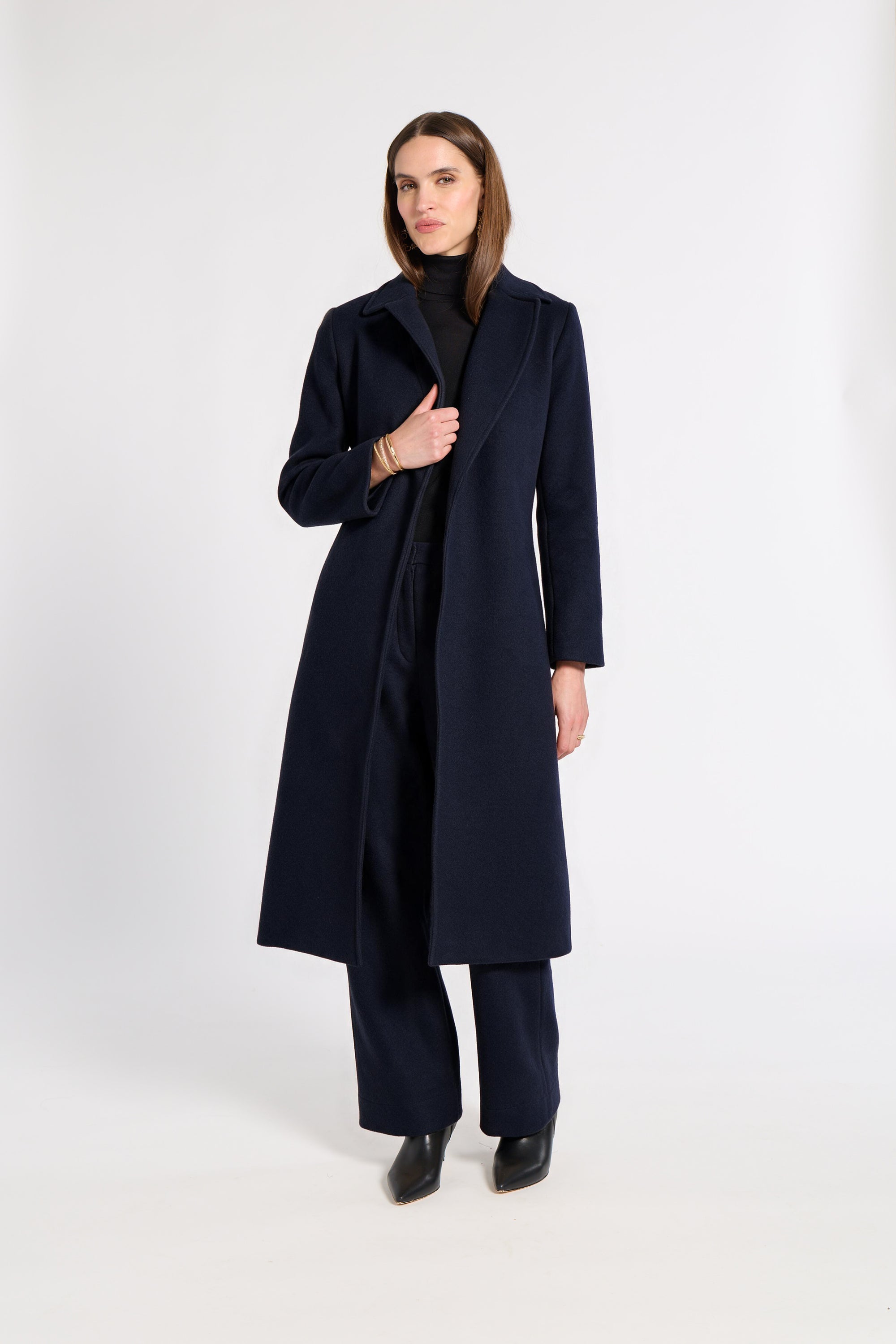 A woman modeling a long navy blue cashmere coat over a black turtleneck and matching navy trousers, standing against a plain white studio background.