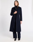 A woman modeling a long navy blue cashmere coat over a black turtleneck and matching navy trousers, standing against a plain white studio background.