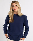 A blonde woman wearing a navy blue knit crewneck sweater and cream trousers, standing against a light gray studio background.