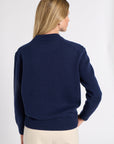 A woman shown from the back wearing a navy blue knit sweater and cream trousers against a plain light gray studio background.