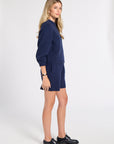 A woman modeling a navy blue knit lounge set consisting of a long-sleeve sweatshirt and matching shorts, paired with black leather loafers, against a plain white studio background.