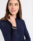 A woman wearing a navy blue long-sleeve button-up shirt with a high mock neck, styled with gold bangles and a gold watch, set against a plain light gray background.