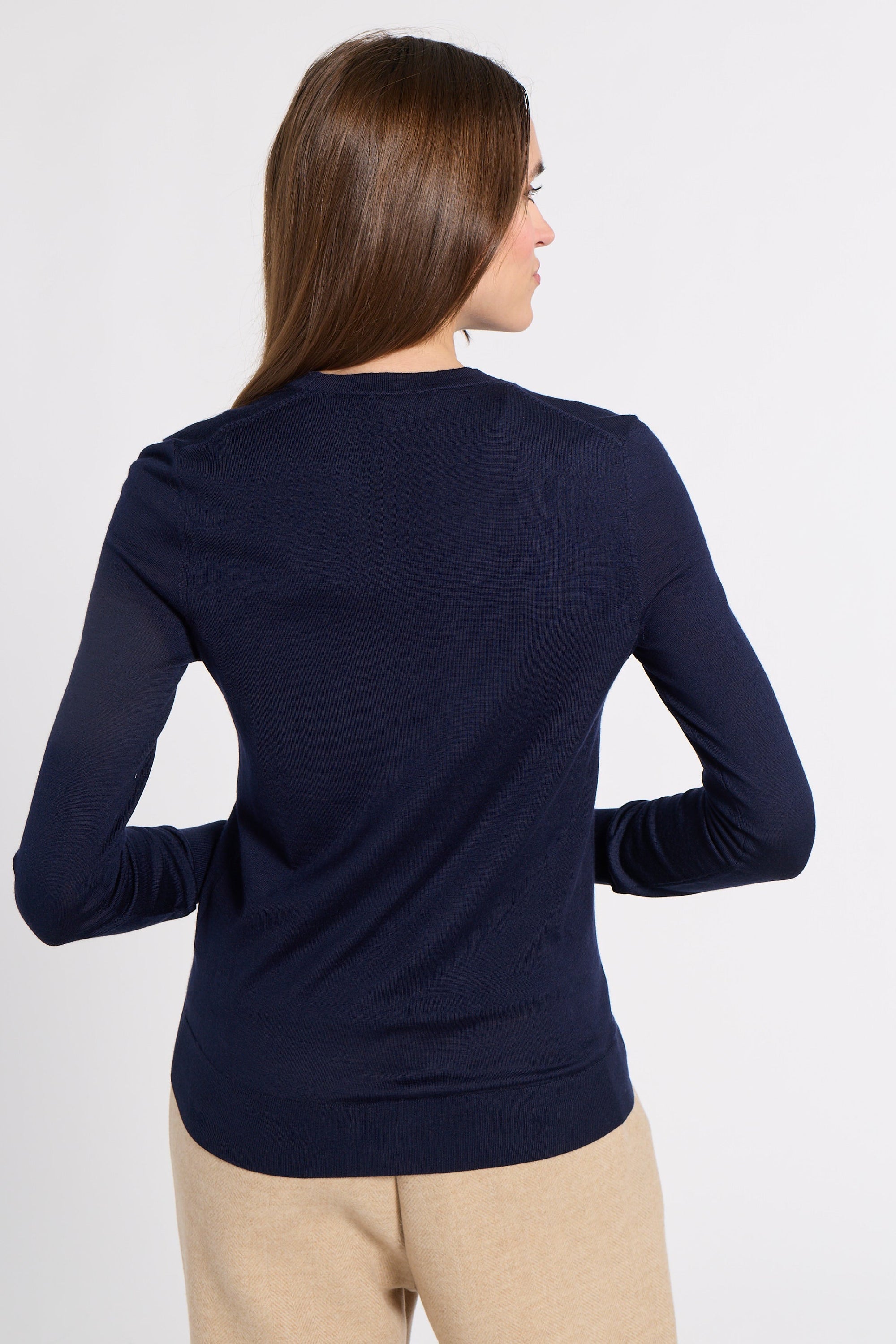 A woman shown from the back wearing a navy blue long-sleeve knit top and beige pants against a plain light gray studio background.