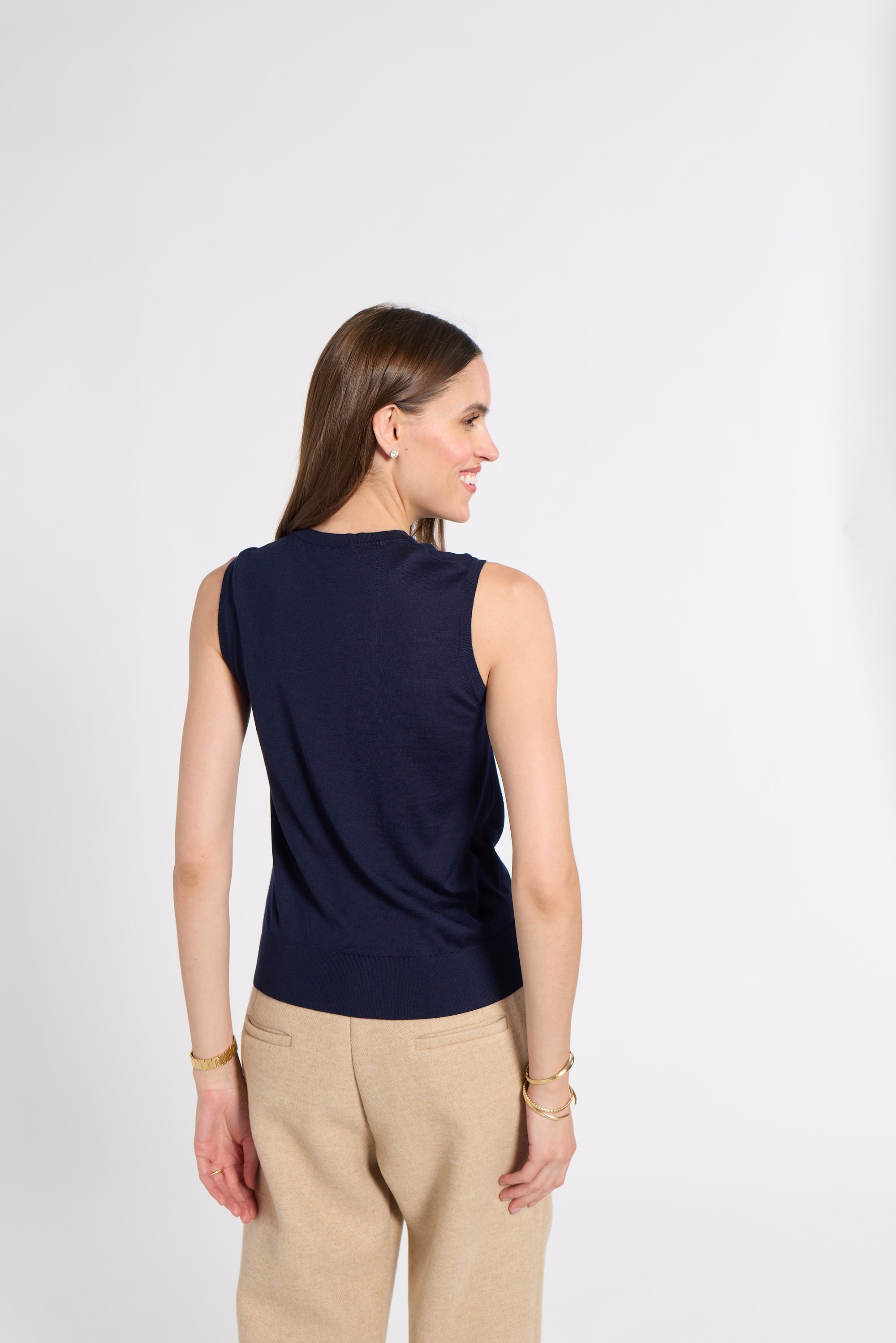 A woman shown from the back wearing a sleeveless navy top and beige pants, standing against a plain light gray studio background.