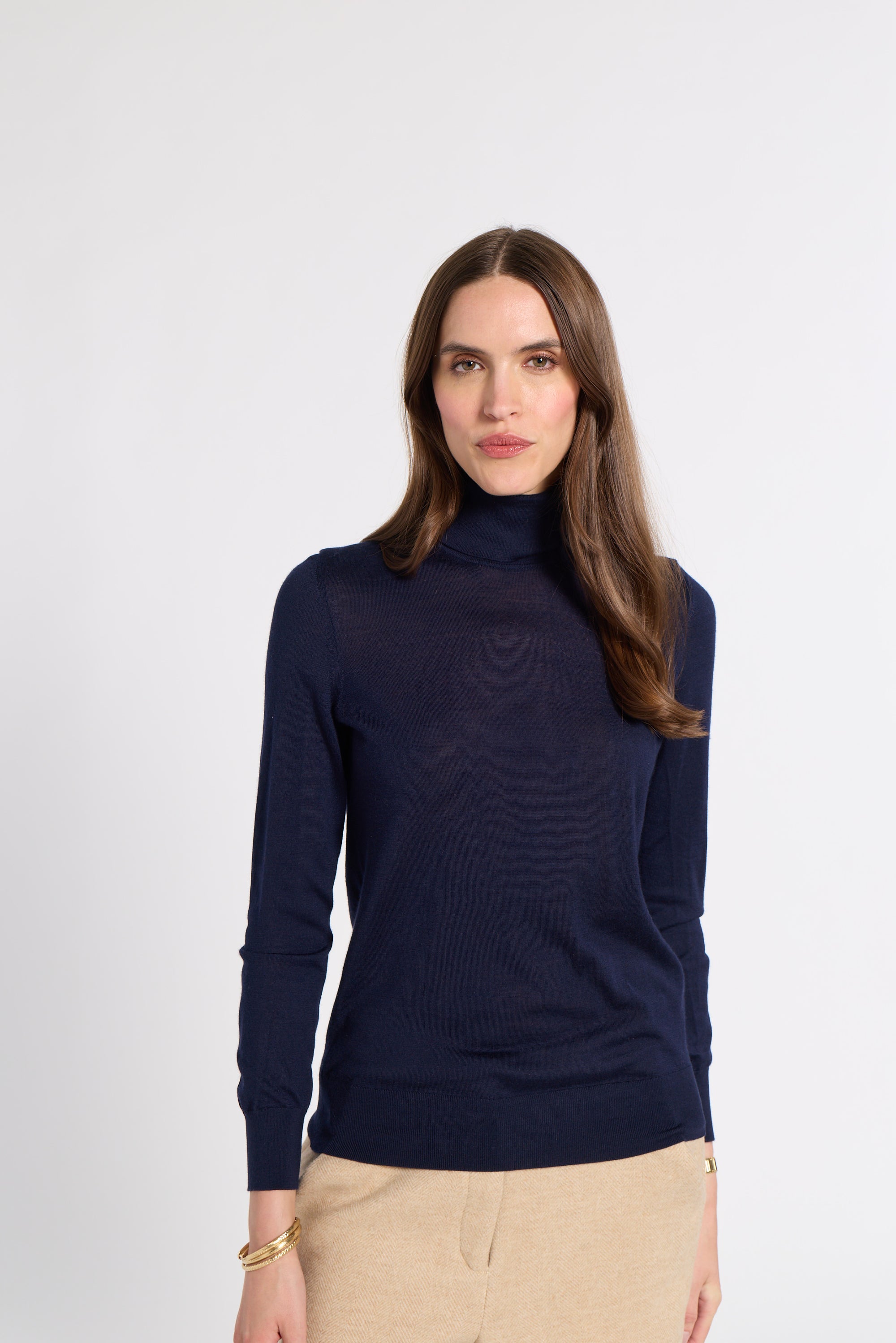 A woman modeling a navy blue long-sleeve turtleneck sweater, paired with beige trousers, against a plain light gray background.