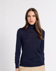 A woman modeling a navy blue long-sleeve turtleneck sweater, paired with beige trousers, against a plain light gray background.