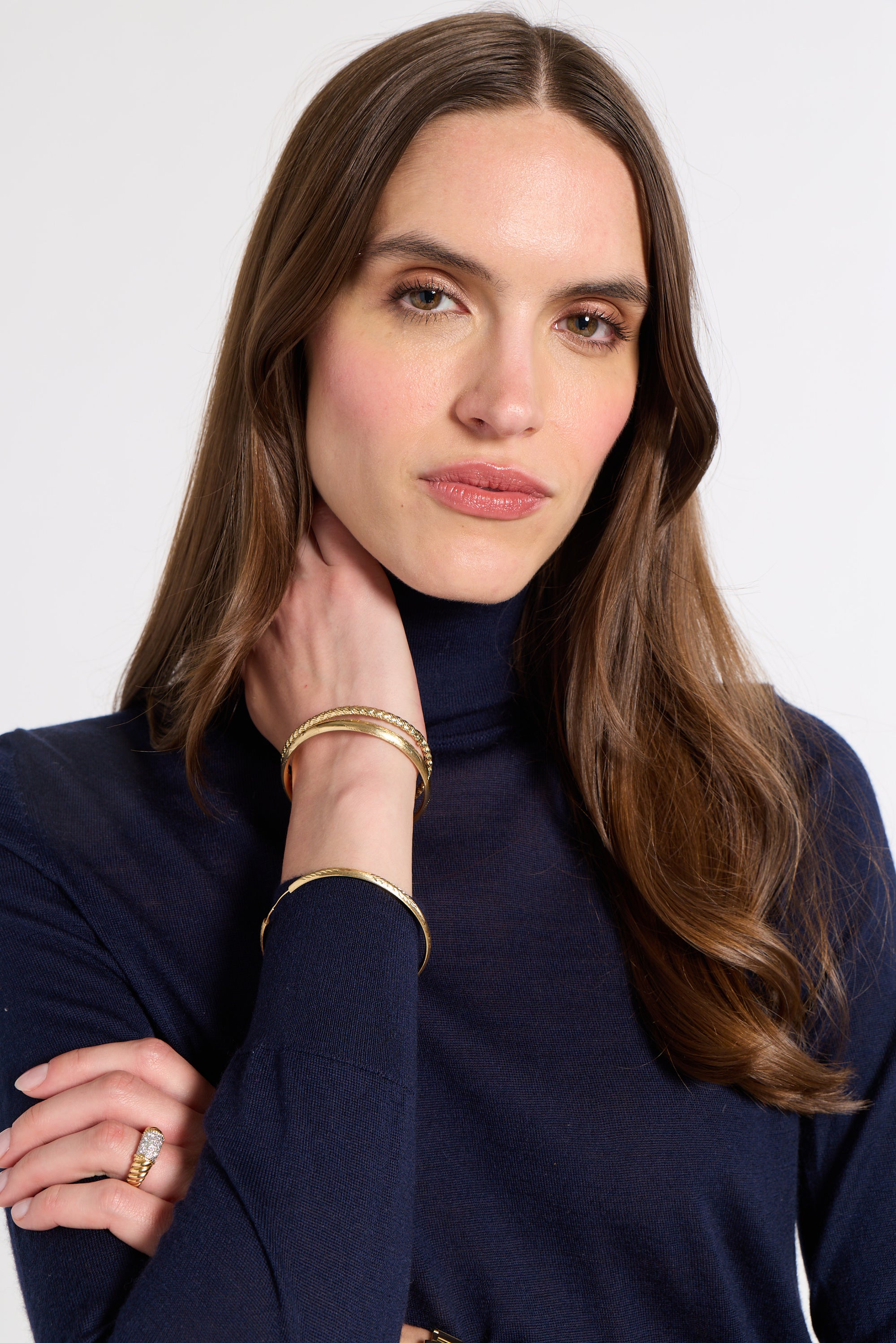 A woman with long brown hair wearing a navy blue turtleneck top and gold bangles, posing against a light gray background.