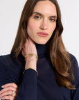 A woman with long brown hair wearing a navy blue turtleneck top and gold bangles, posing against a light gray background.