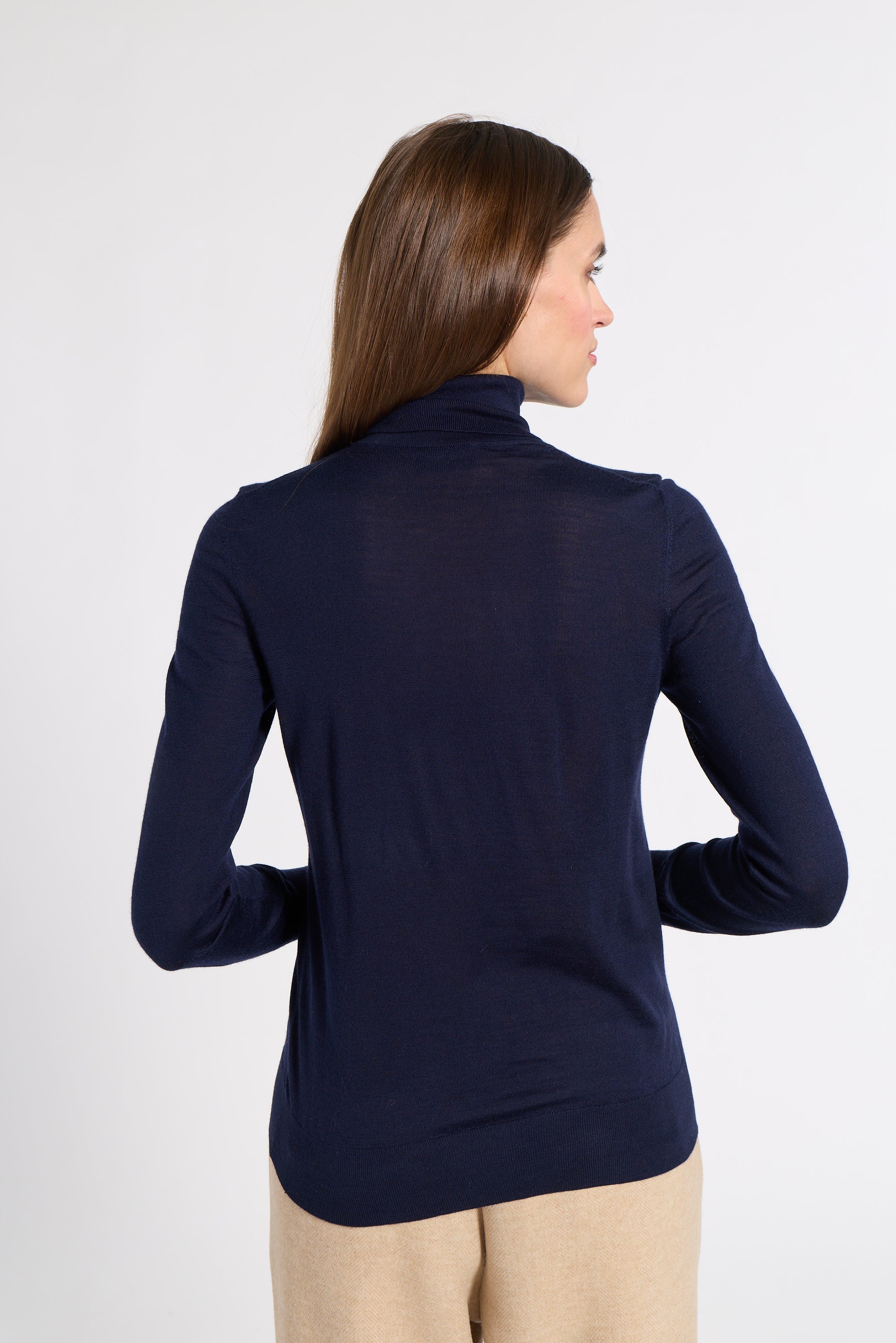 A woman shown from the back wearing a dark navy long-sleeve turtleneck top and beige pants, photographed against a light gray studio background.