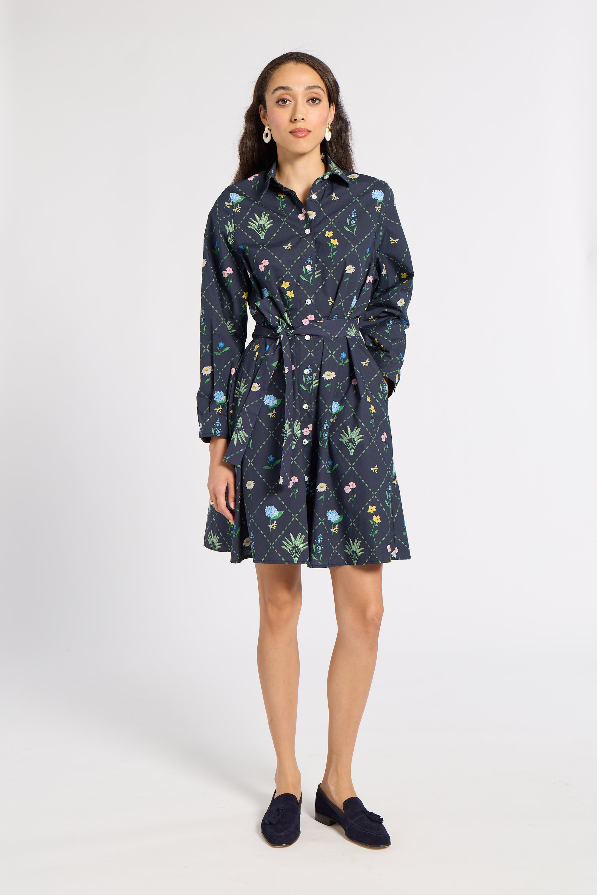 A woman modeling a dark navy blue long-sleeve shirt dress with a colorful floral and botanical print, featuring a button-up front, collar, and self-tie waist belt, paired with dark loafers against a light gray studio background.