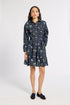 A woman modeling a dark navy blue long-sleeve shirt dress with a colorful floral and botanical print, featuring a button-up front, collar, and self-tie waist belt, paired with dark loafers against a light gray studio background.