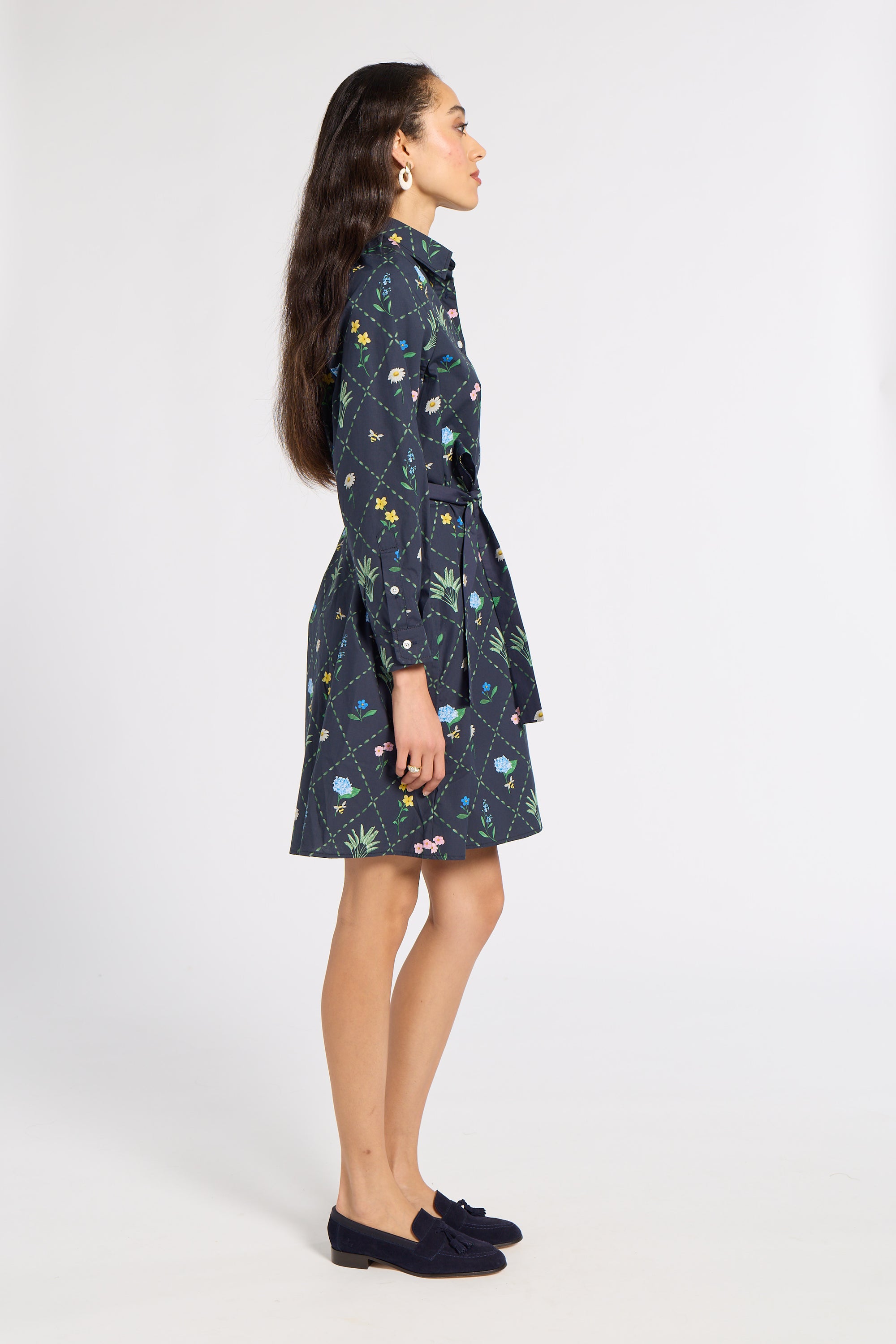 A woman stands in profile wearing a dark navy long-sleeve mini dress with a colorful floral and butterfly print, paired with dark navy loafers, against a plain light gray studio background.