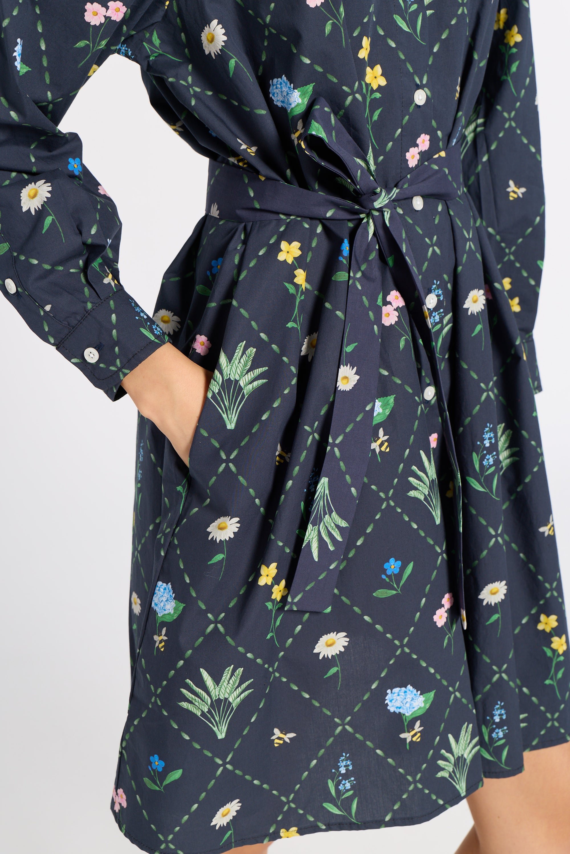 A close-up of a navy blue long-sleeve dress with a colorful floral and green leaf print, featuring a tie waist and side pockets.