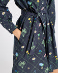 A close-up of a navy blue long-sleeve dress with a colorful floral and green leaf print, featuring a tie waist and side pockets.