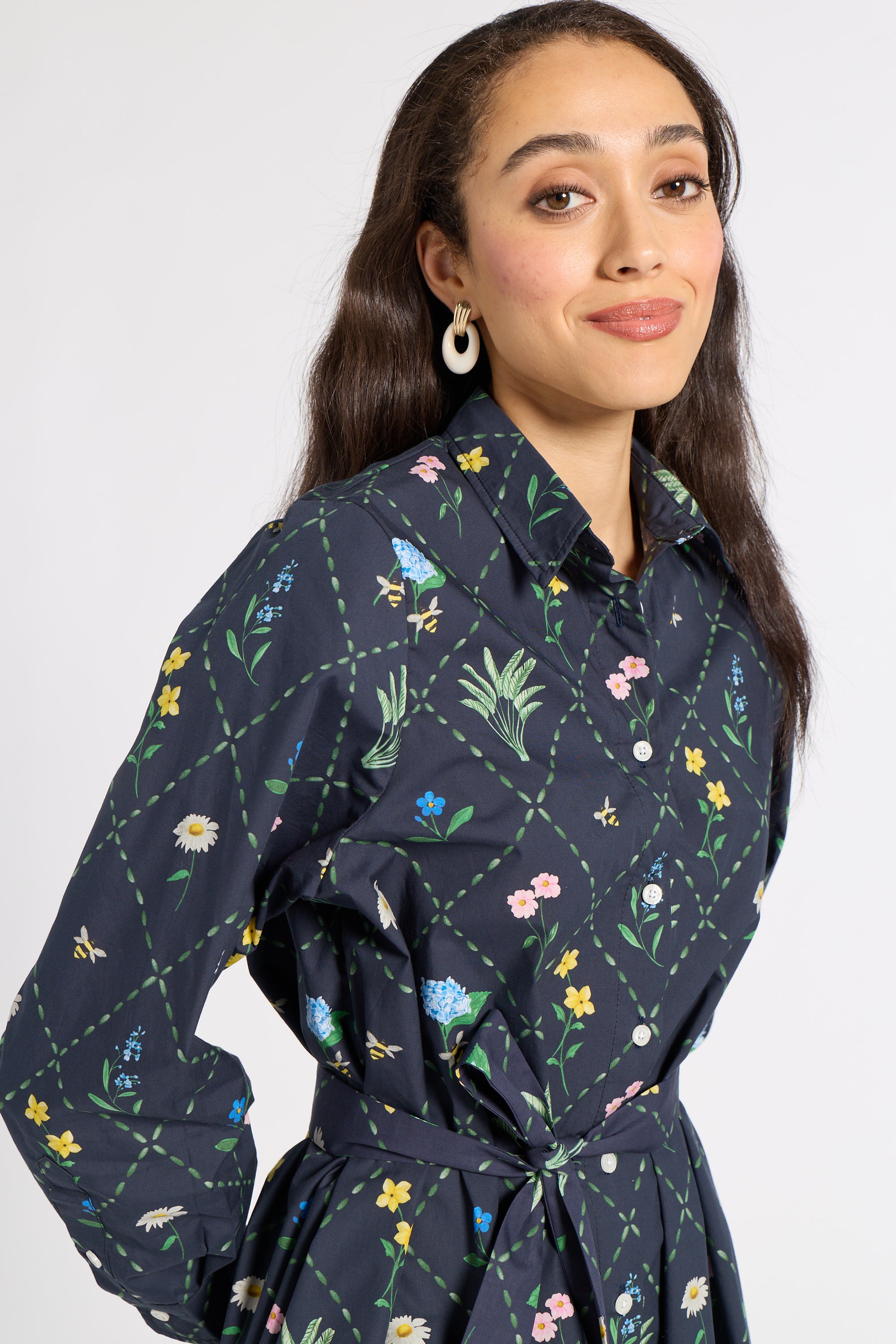 A woman wearing a dark navy long-sleeve button-up shirt with a colorful floral and insect print, featuring a matching waist tie belt, set against a plain light gray background.