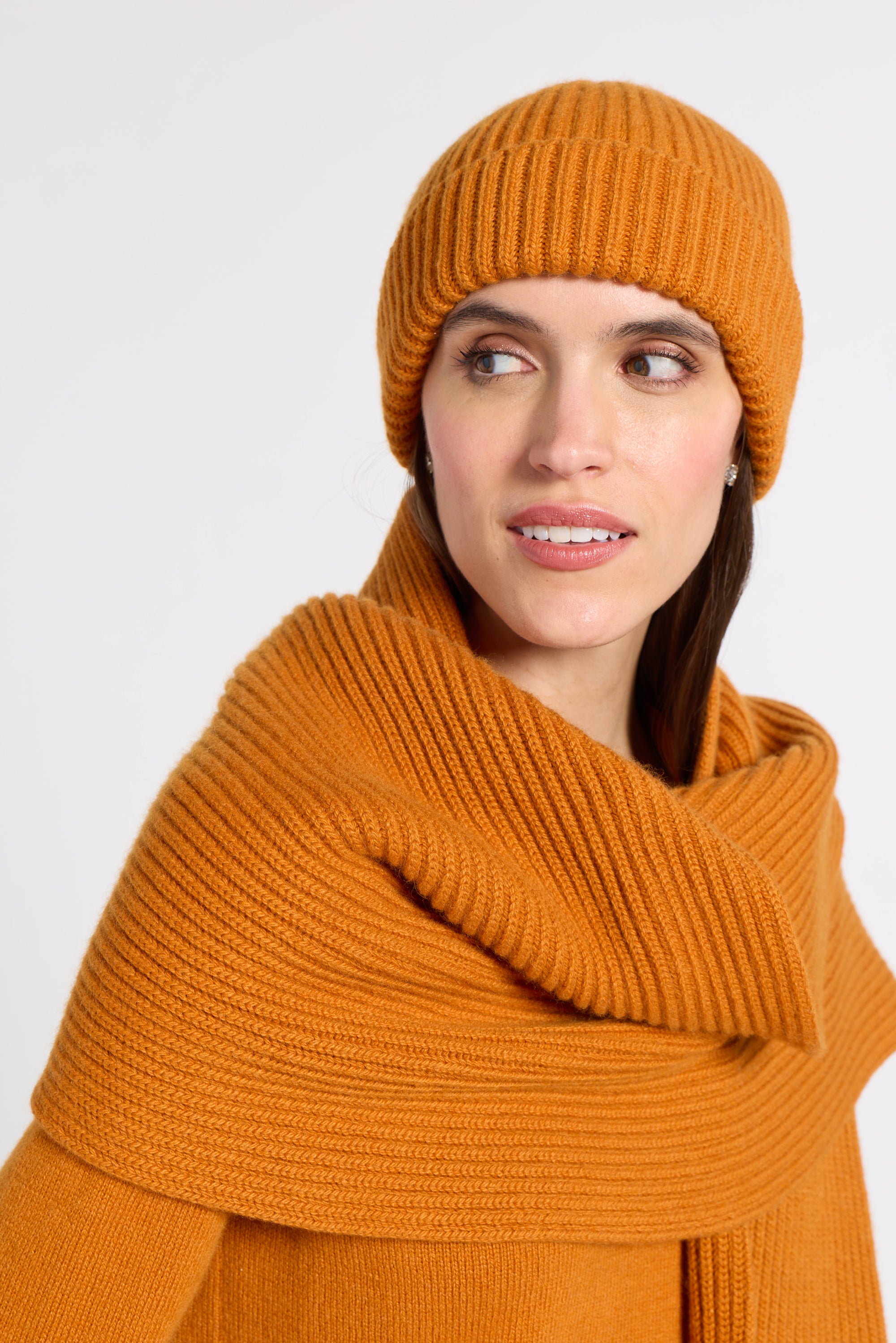 A woman wearing a matching ribbed knit beanie and thick scarf in a warm orange-brown tone, photographed against a clean light gray studio background.