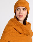 A woman wearing a matching ribbed cashmere beanie and scarf in a warm orange-brown tone, photographed against a clean light gray studio background.