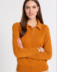 A woman with long brown hair wearing a mustard orange knit polo sweater and beige trousers, standing with arms crossed against a light gray studio background.