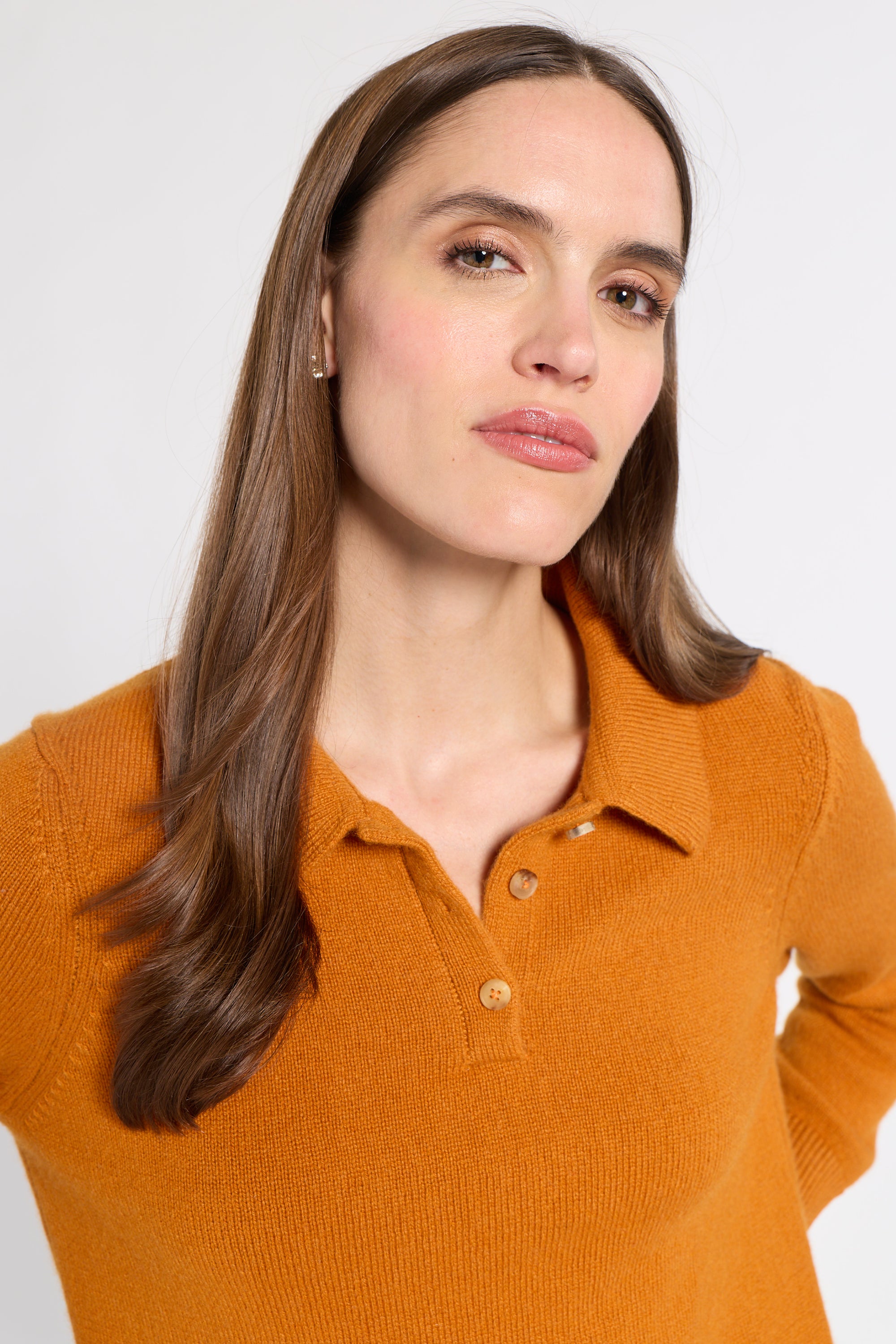 A woman with long brown hair wearing a mustard orange knit polo shirt, photographed against a clean light gray studio background.