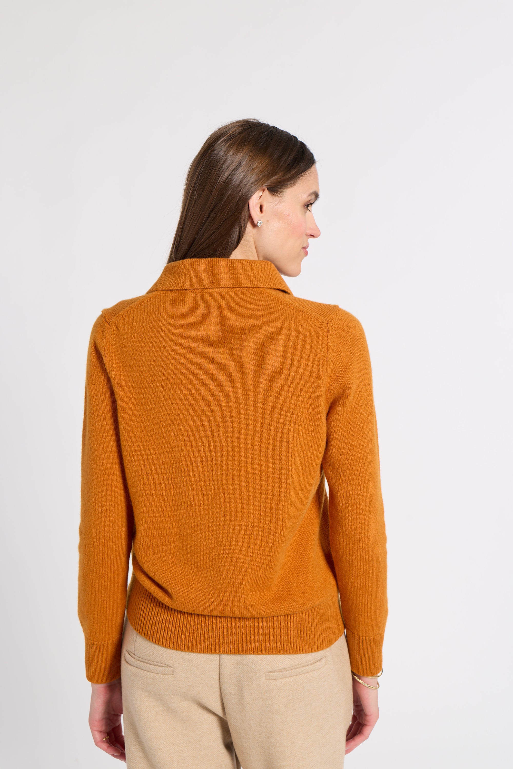 A woman shown from the back wearing a camel-colored knit sweater and beige trousers, photographed against a plain light gray studio background.