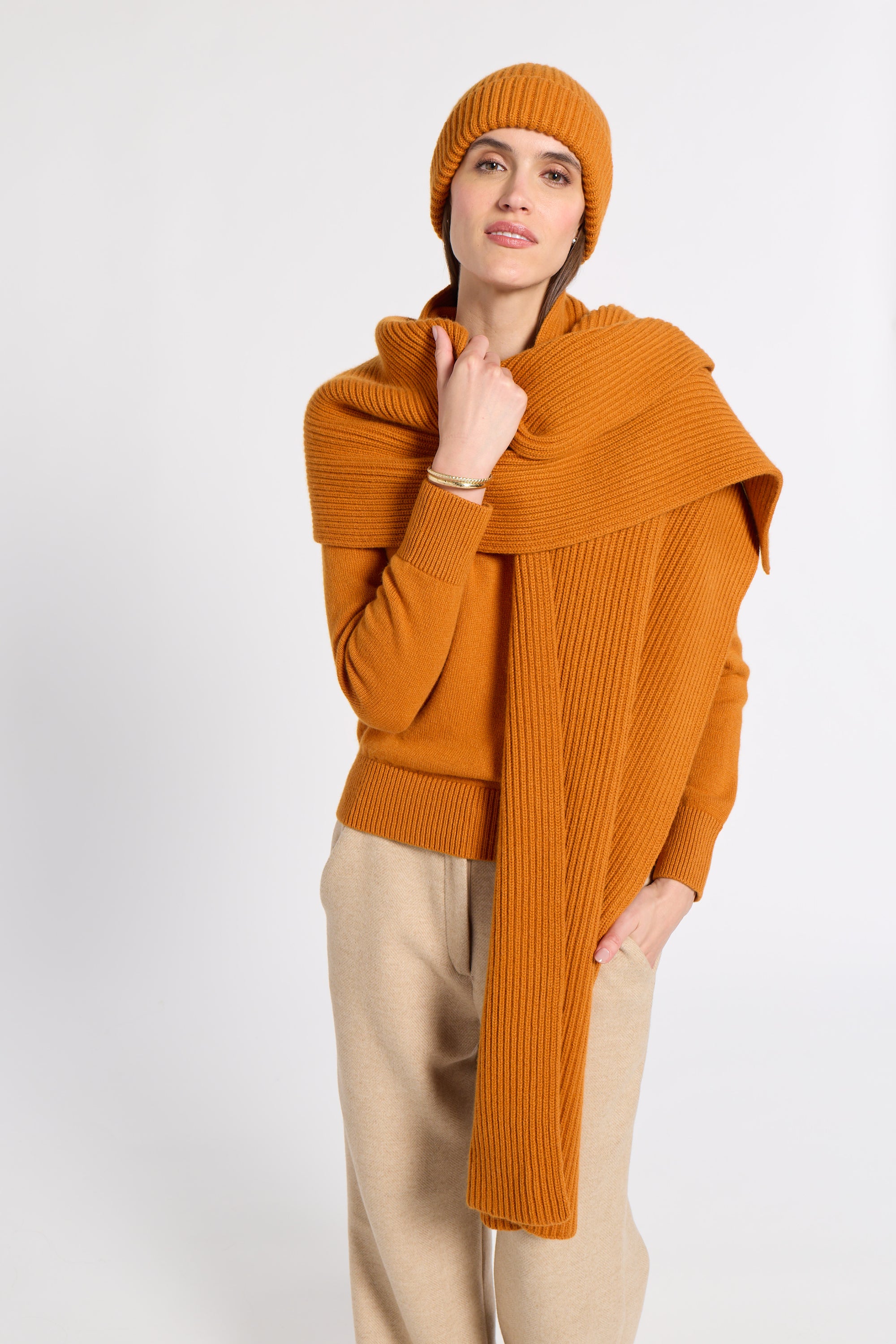 A woman modeling a warm orange ribbed knit set, including a beanie, a large draped scarf, and a matching sweater, paired with beige trousers, against a plain light gray background.