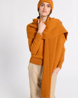 A woman modeling a warm orange ribbed knit set, including a beanie, a large draped scarf, and a matching sweater, paired with beige trousers, against a plain light gray background.