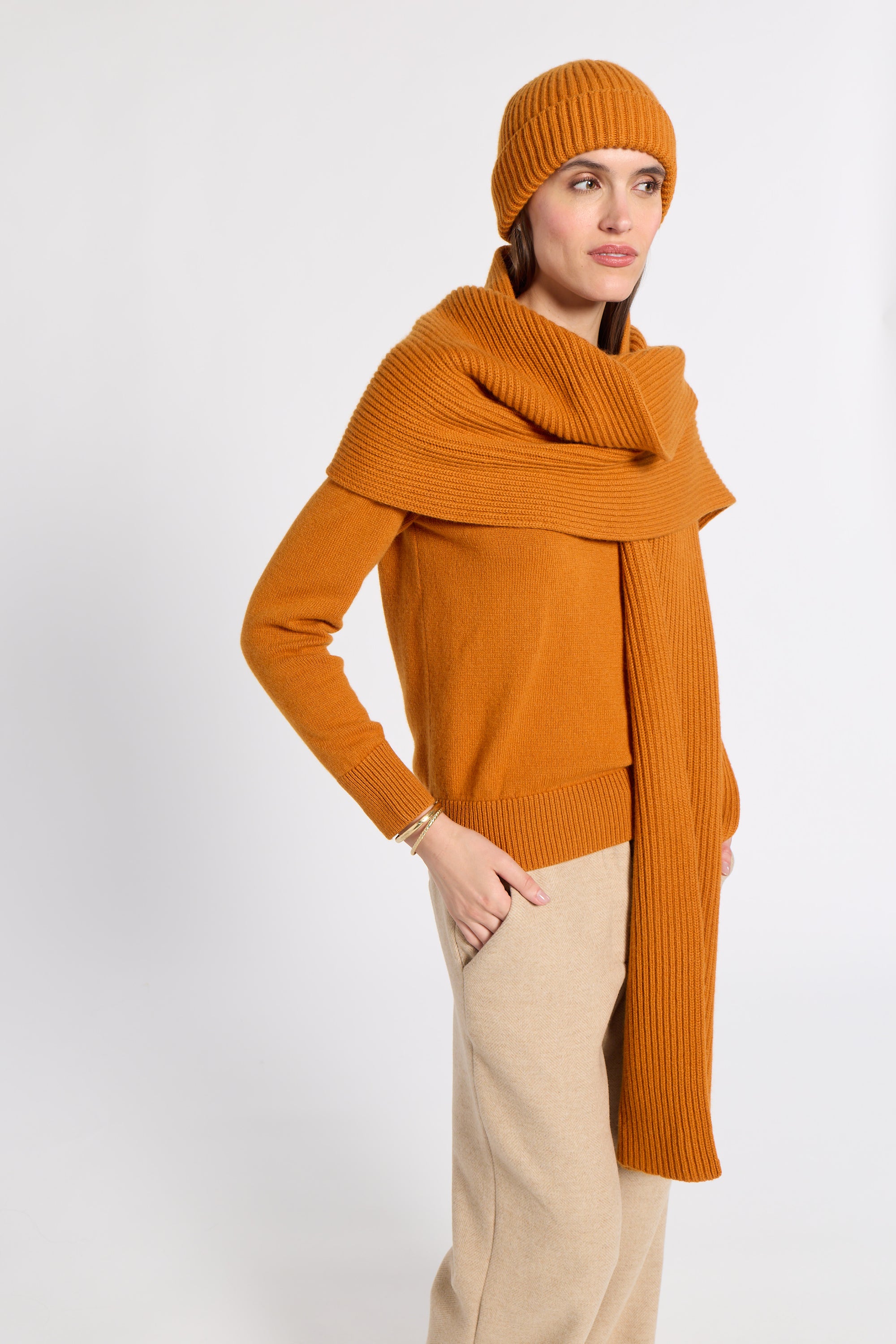 A woman modeling a warm orange ribbed knit sweater, matching beanie, and a wide draped scarf, paired with beige trousers, against a plain light gray studio background.