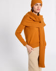 A woman modeling a warm orange ribbed knit sweater, matching beanie, and a wide draped scarf, paired with beige trousers, against a plain light gray studio background.