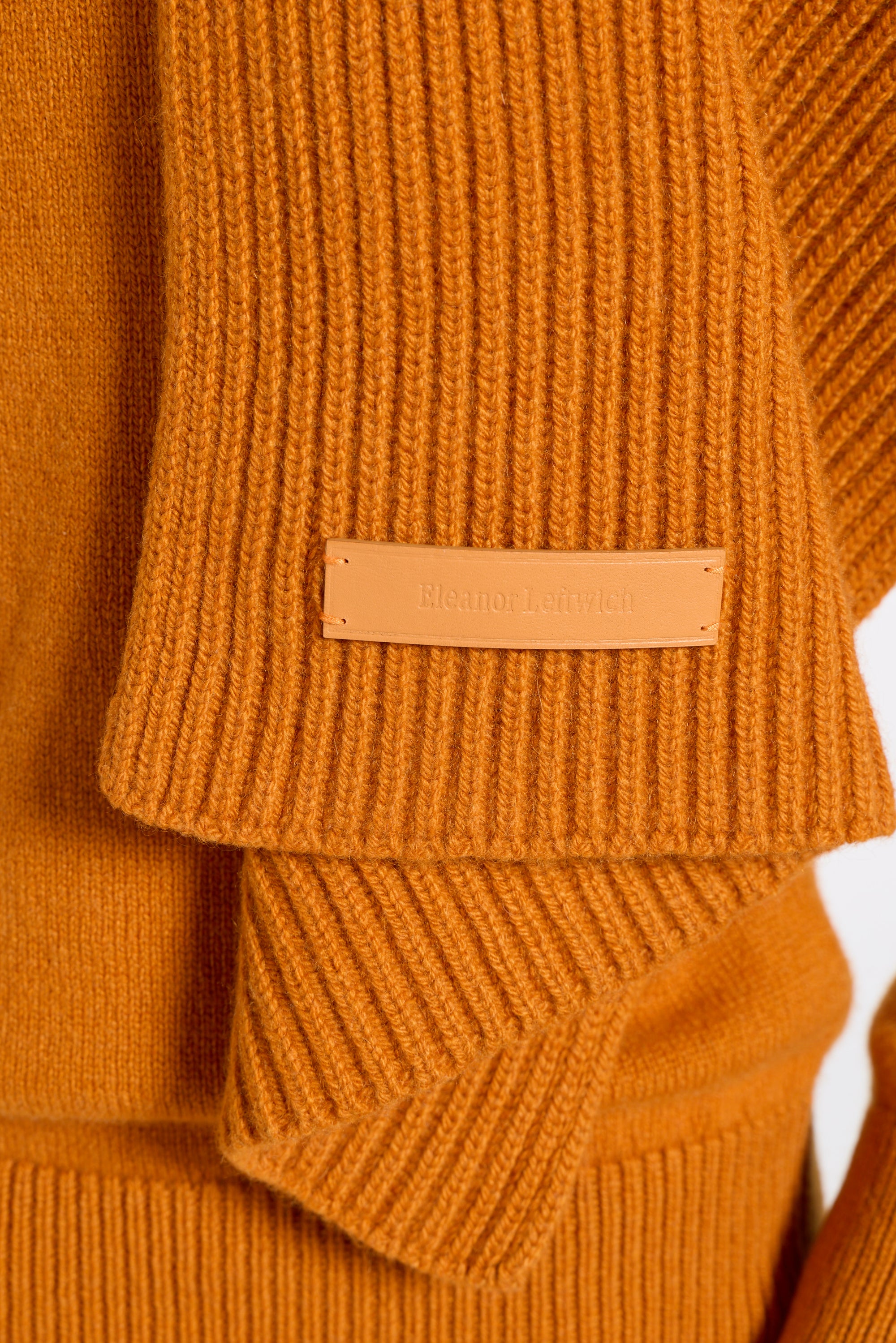 Close-up of a ribbed knit sweater in a warm camel orange color, showing a tan leather brand patch on the sleeve with the text 'BONMALI' visible.