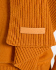 Close-up of a ribbed knit sweater in a warm camel orange color, showing a tan leather brand patch on the sleeve with the text 'BONMALI' visible.