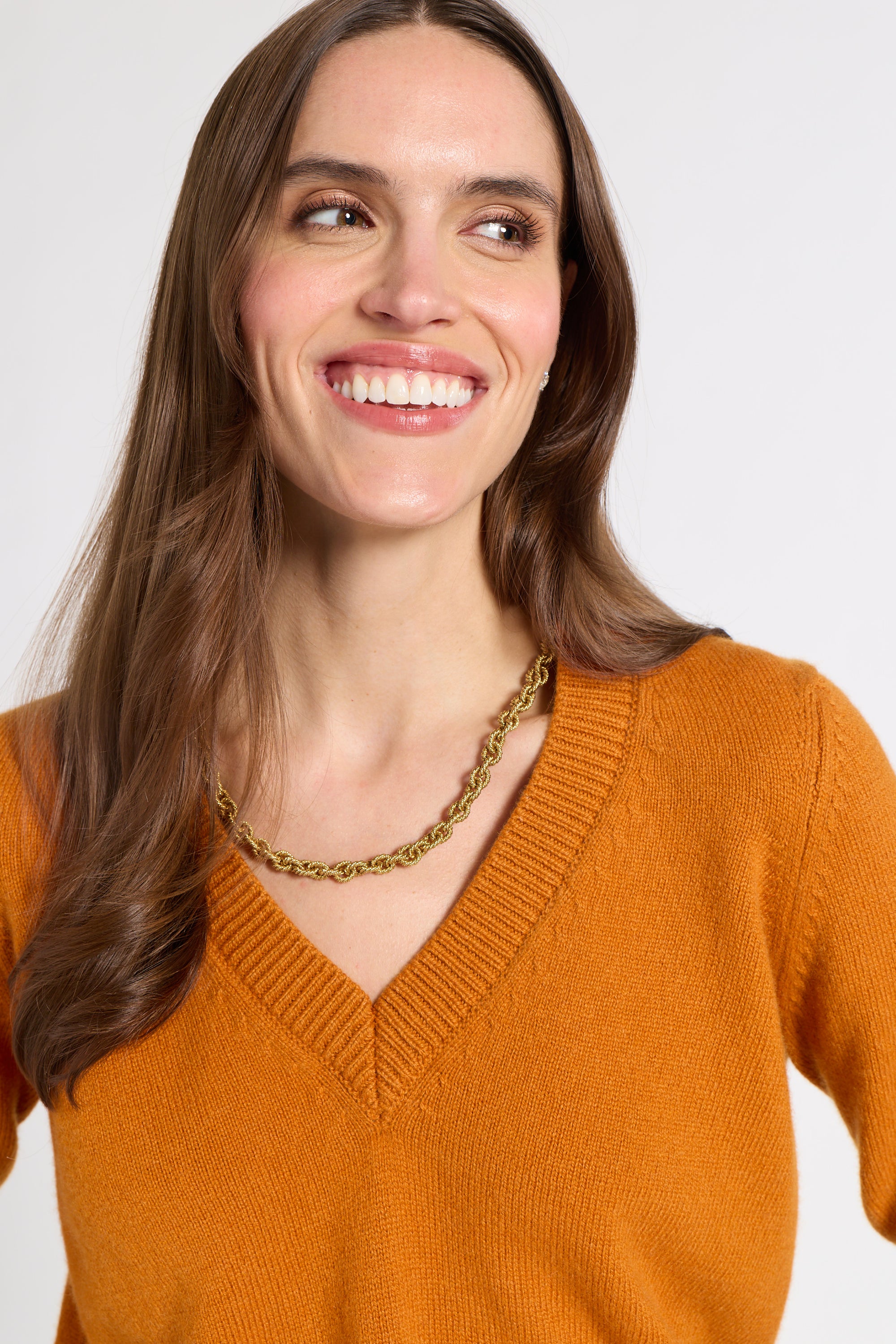 A smiling woman with long brown hair wearing a mustard orange V-neck knit sweater and a gold chain necklace, photographed against a light gray studio background.