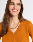 A smiling woman with long brown hair wearing a mustard orange V-neck knit sweater and a gold chain necklace, photographed against a light gray studio background.