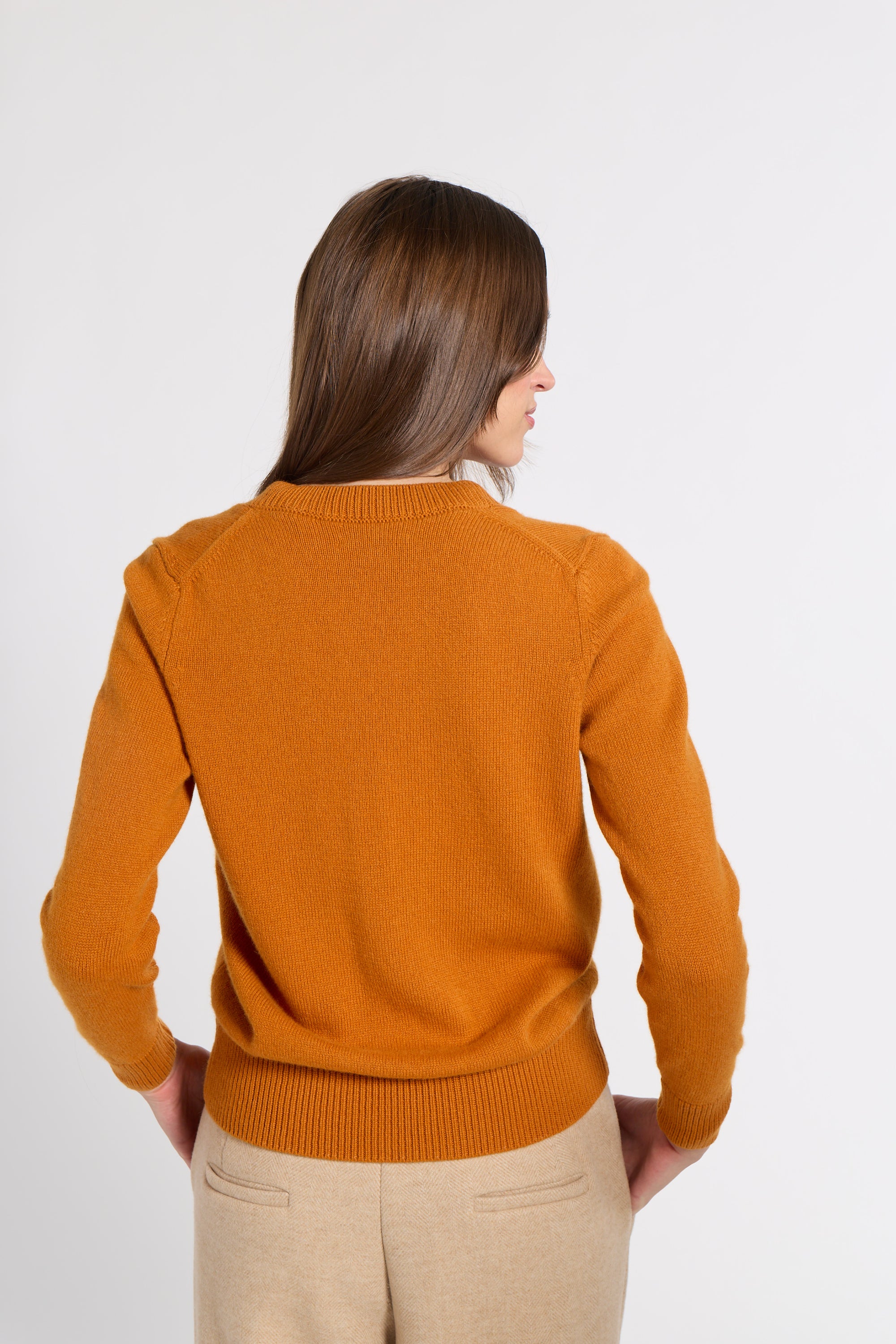 A woman viewed from behind wearing a mustard orange knit sweater and beige trousers against a plain light gray studio background.