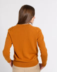 A woman viewed from behind wearing a mustard orange knit sweater and beige trousers against a plain light gray studio background.