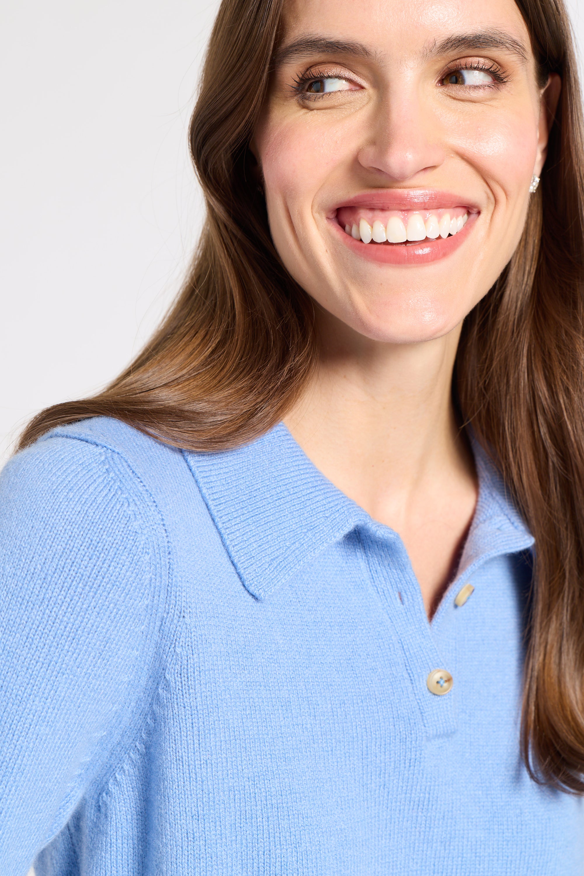 A close-up of a woman wearing a light blue knit polo shirt, smiling and looking to the side against a plain light gray background.