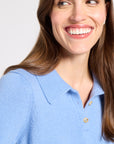 A close-up of a woman wearing a light blue knit polo shirt, smiling and looking to the side against a plain light gray background.