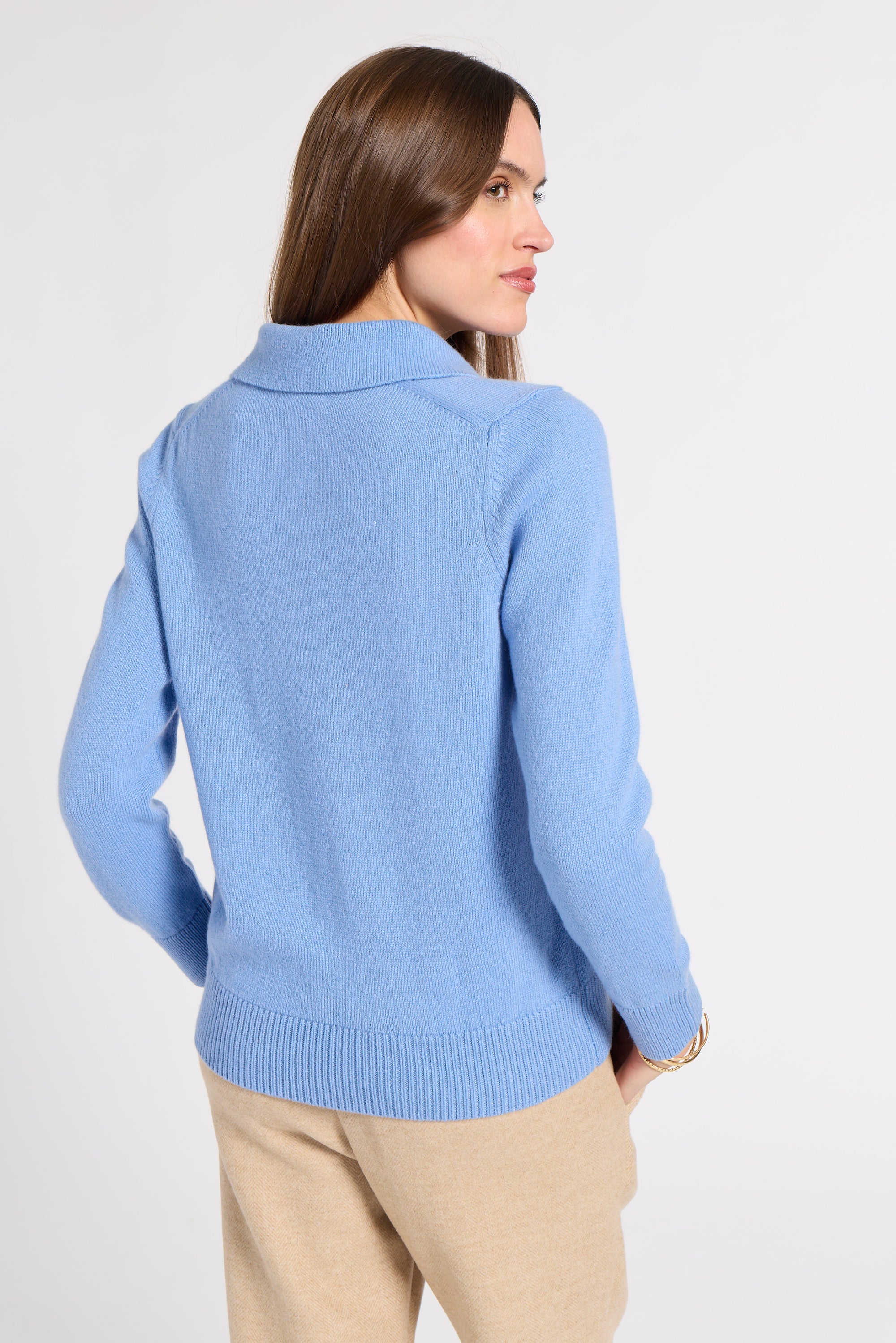 A woman shown from the back wearing a light blue knit sweater and beige pants against a plain light gray studio background.