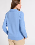 A woman shown from the back wearing a light blue knit sweater and beige pants against a plain light gray studio background.