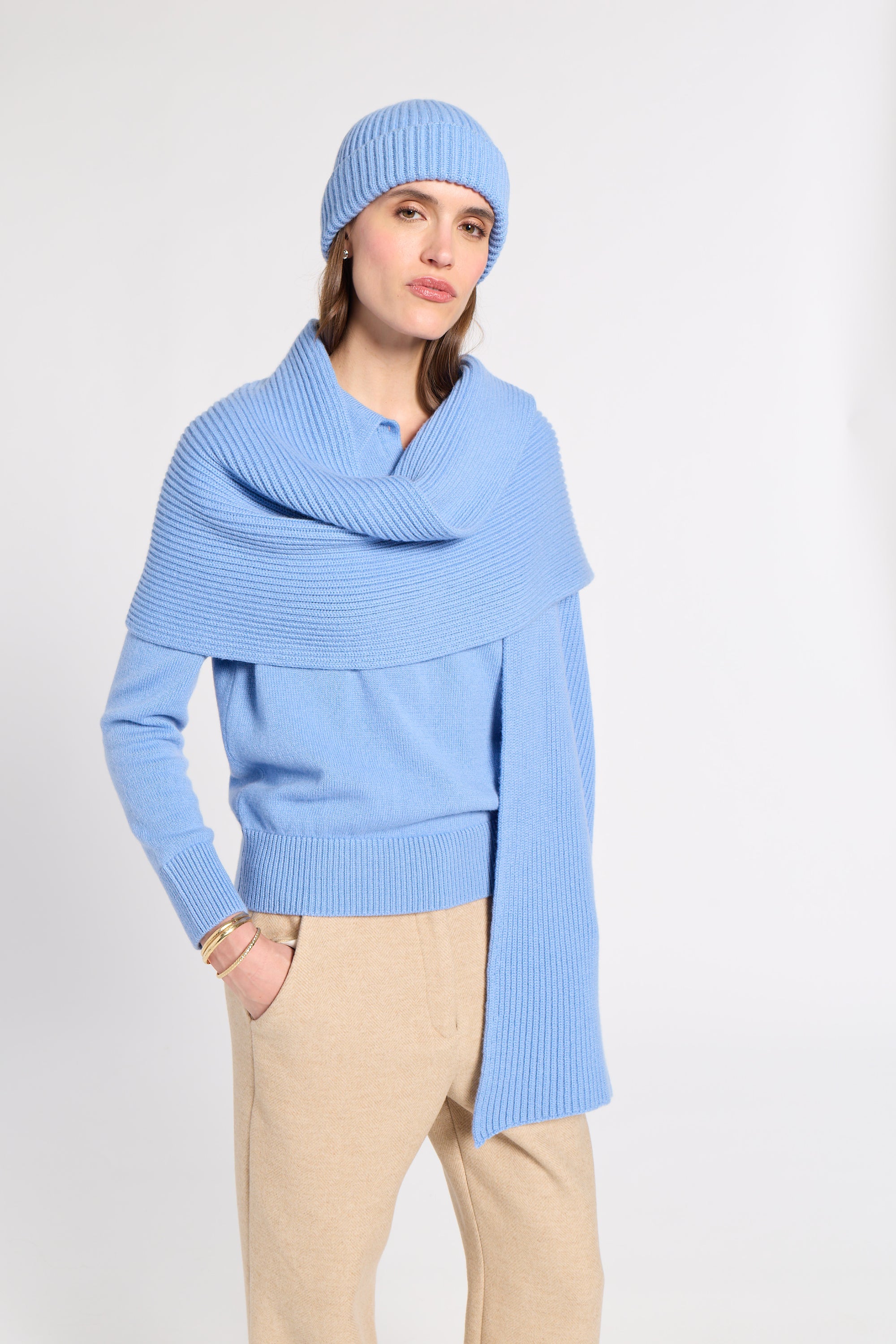 A woman modeling a light blue ribbed knit beanie, matching wrap shawl, and light blue knit sweater, paired with beige trousers, against a plain light gray studio background.