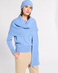A woman modeling a light blue ribbed knit beanie, matching wrap shawl, and light blue knit sweater, paired with beige trousers, against a plain light gray studio background.