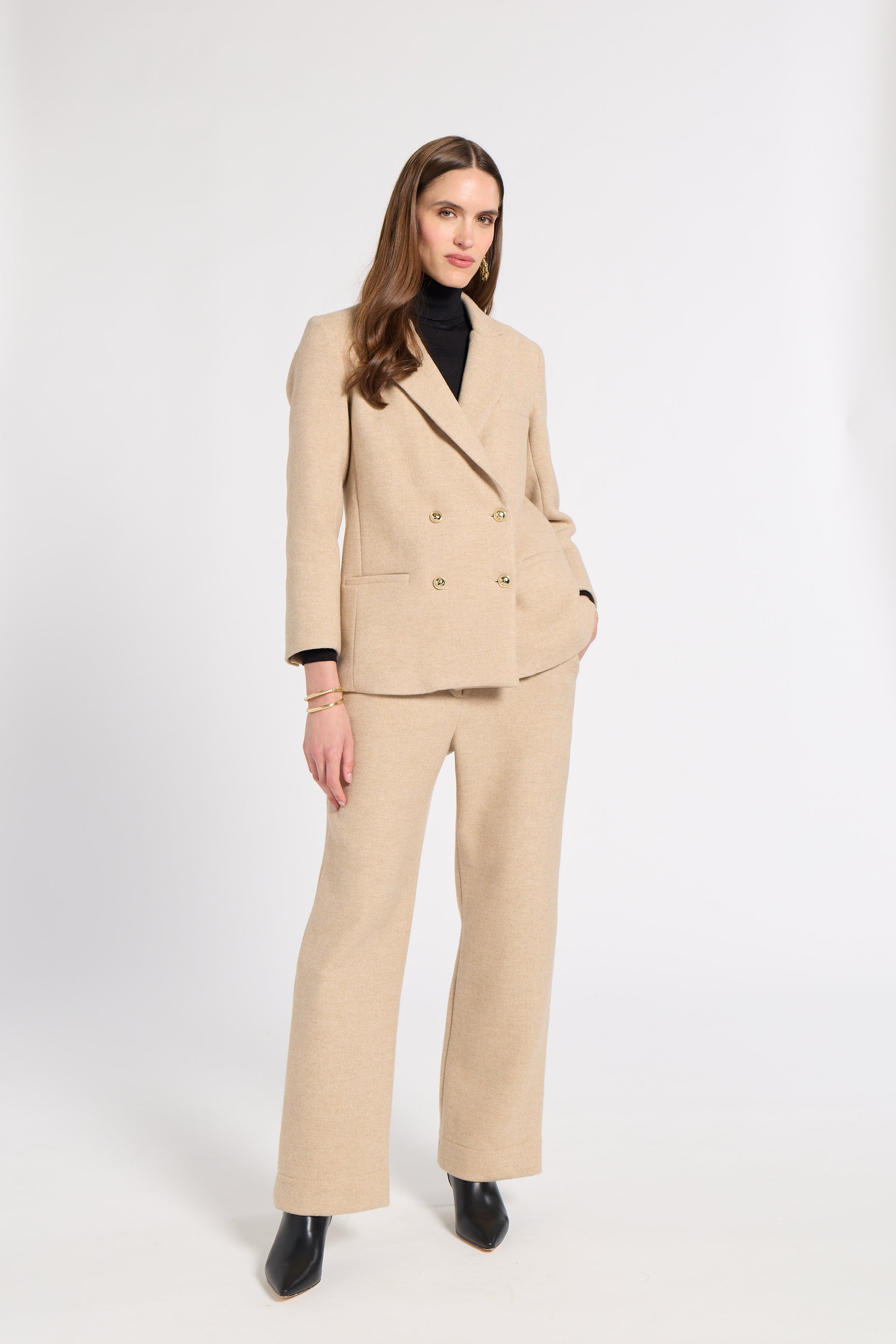 A woman modeling a beige double-breasted suit with wide-leg trousers, worn over a black turtleneck and black pointed shoes, against a plain white studio background.