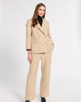 A woman modeling a beige double-breasted suit with wide-leg trousers, worn over a black turtleneck and black pointed shoes, against a plain white studio background.