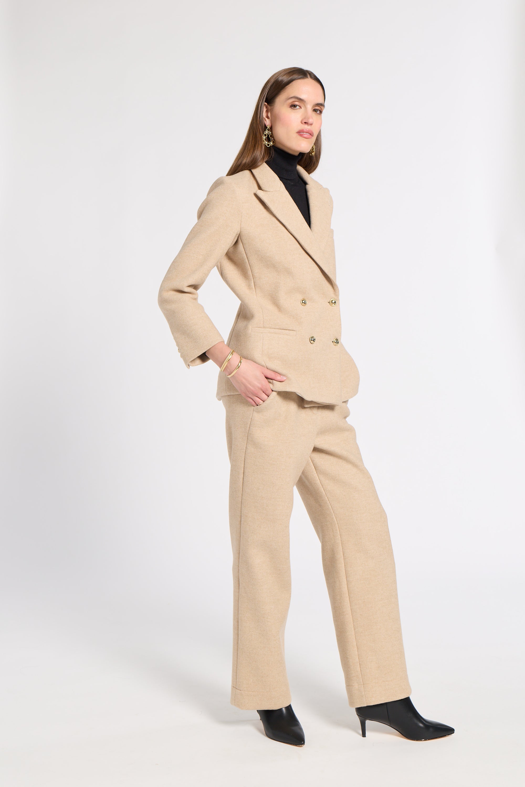 A woman models a beige double-breasted blazer and matching wide-leg trousers set, styled with a black turtleneck and black pointed-toe heels against a plain white studio background.