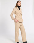 A woman models a beige double-breasted blazer and matching wide-leg trousers set, styled with a black turtleneck and black pointed-toe heels against a plain white studio background.
