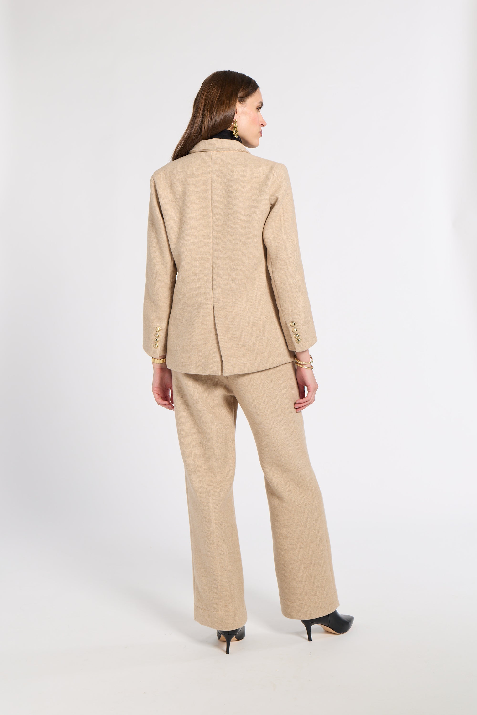 A woman is shown from the back wearing a beige tailored suit consisting of a blazer and wide-leg trousers, paired with black pointed-toe heels, against a plain light gray studio background.