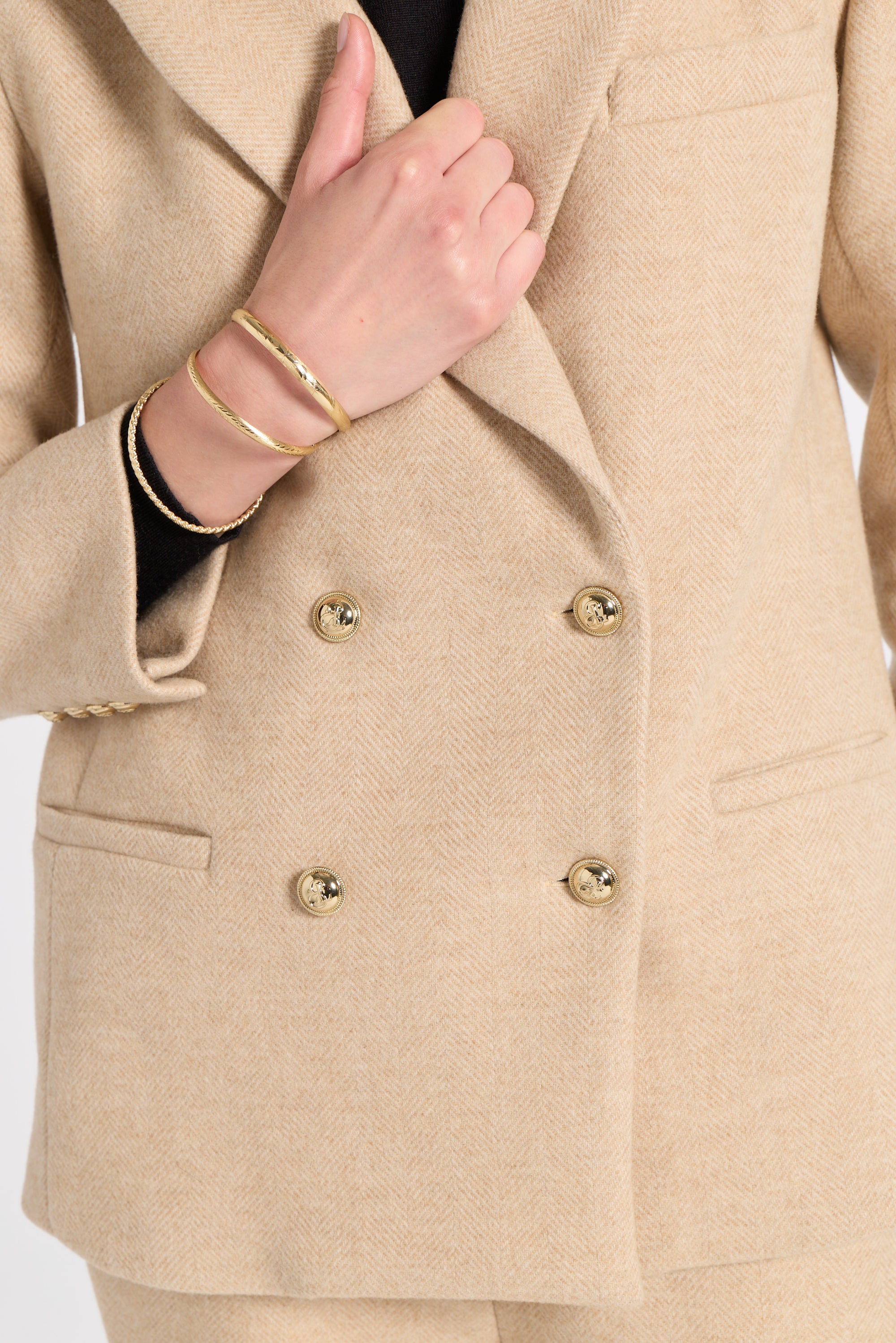 Close-up of a beige double-breasted blazer with gold buttons, worn by a model whose hand and wrist with gold bracelets are visible.